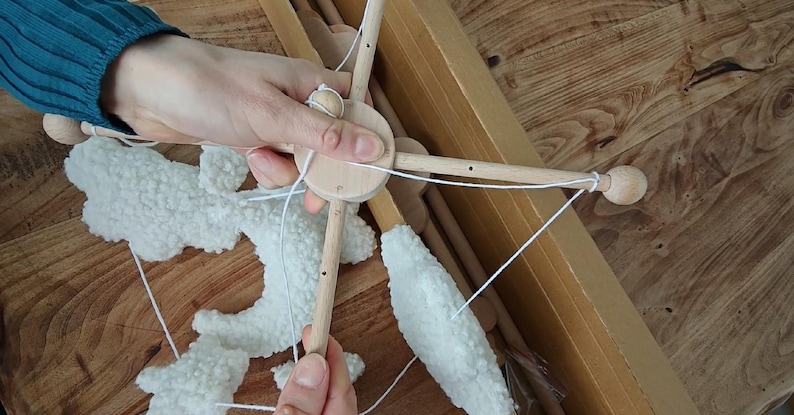 May include: A wooden baby mobile frame featuring a central circular hub with four extending arms, each ending in a small wooden sphere. White, textured, cloud-shaped fabric pieces are suspended from the frame by thin white strings.