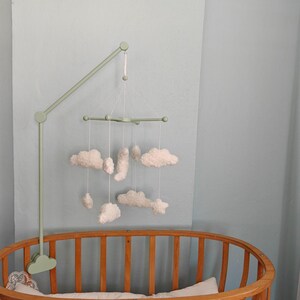 May include: A sage green mobile arm attached to a wooden crib, featuring a cross-shaped hanger suspended by white strings. The mobile displays several white, plush, cloud-shaped ornaments of varying sizes hanging at different heights.