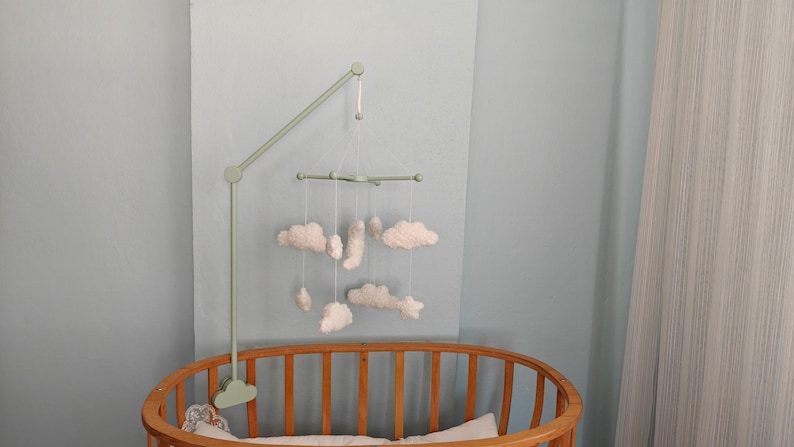 May include: A sage green mobile arm attached to a wooden crib, featuring a cross-shaped hanger suspended by white strings. The mobile displays several white, plush, cloud-shaped ornaments of varying sizes hanging at different heights.