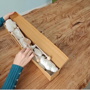 May include: A long, rectangular cardboard box containing a decorative garland made of white, fluffy, cloud-like shapes connected by thin, light-colored wooden rods and white string. The box is open, revealing the contents arranged in a linear fashion.