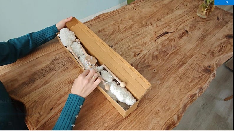 May include: A long, rectangular cardboard box containing a decorative garland made of white, fluffy, cloud-like shapes connected by thin, light-colored wooden rods and white string. The box is open, revealing the contents arranged in a linear fashion.