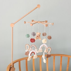 May include: A wooden baby mobile with a teddy bear, pom-poms, and rainbow-shaped crocheted decorations hanging from it. The mobile is attached to a wooden arm that extends from a crib.