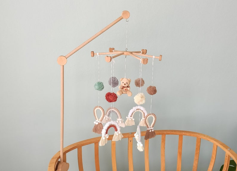 May include: A wooden baby mobile with a teddy bear, pom-poms, and rainbow-shaped crocheted decorations hanging from it. The mobile is attached to a wooden arm that extends from a crib.