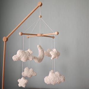 May include: A wooden baby mobile featuring a crescent moon, stars, and fluffy cloud shapes. The mobile is suspended from a wooden frame with a natural finish. The clouds and celestial shapes are white and textured, creating a soft, whimsical aesthetic.