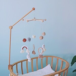 May include: A wooden crib mobile arm attached to a light wood oval crib. The mobile features hanging white strings with soft pom-poms in shades of rust, gray, and cream, alongside macrame rainbow and loop shapes in neutral earth tones.