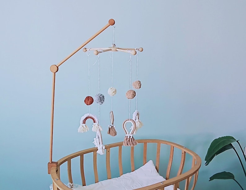 May include: A wooden crib mobile arm attached to a light wood oval crib. The mobile features hanging white strings with soft pom-poms in shades of rust, gray, and cream, alongside macrame rainbow and loop shapes in neutral earth tones.