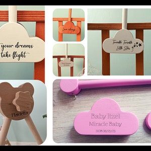 May include: A set of wooden baby mobile parts with cloud-shaped pieces that can be personalized with text. The clouds are painted in white, pink, and light brown. The text on the clouds includes "Let your dreams take flight", "Love always", "Twinkle Twinkle Little Star", "Baby Itzel Miracle Baby DOB 08/25/23", and "Flavietta".