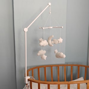 May include: A white crib mobile arm attached to a wooden oval crib, featuring hanging white plush ornaments shaped like a crescent moon, stars, and clouds suspended by thin white strings from a central white cross-shaped frame structure.