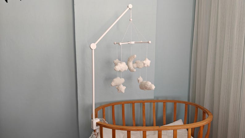 May include: A white crib mobile arm attached to a wooden oval crib, featuring hanging white plush ornaments shaped like a crescent moon, stars, and clouds suspended by thin white strings from a central white cross-shaped frame structure.
