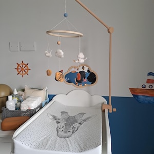 May include: A white changing table with a giraffe print changing pad. A wooden baby mobile hangs above, featuring felt clouds, animals, and a wooden frame. The room is decorated with nautical and animal-themed wall art.