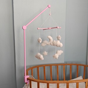 May include: A pink mobile arm attached to a wooden crib, featuring a hanging mobile with white, plush, cloud and star-shaped ornaments suspended by thin white strings from a central pink cross-shaped frame structure.