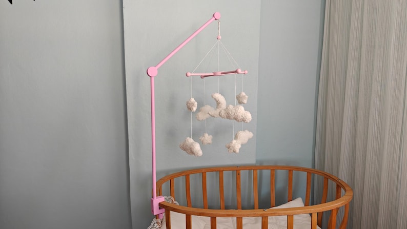 May include: A pink mobile arm attached to a wooden crib, featuring a hanging mobile with white, plush, cloud and star-shaped ornaments suspended by thin white strings from a central pink cross-shaped frame structure.