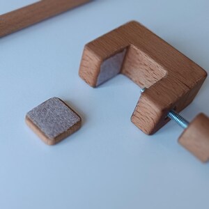 May include: A C-shaped wooden clamp with a threaded metal screw and a matching wooden knob. The clamp features a light gray felt pad on the inner surface. A separate small square wooden piece with a matching felt pad sits nearby, alongside a wooden rod.