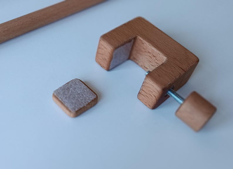May include: A C-shaped wooden clamp with a threaded metal screw and a matching wooden knob. The clamp features a light gray felt pad on the inner surface. A separate small square wooden piece with a matching felt pad sits nearby, alongside a wooden rod.
