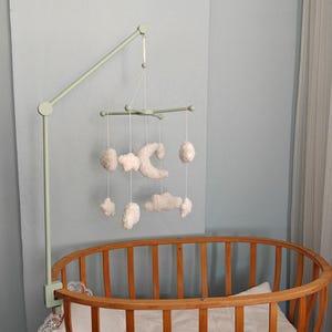 May include: A light green metal arm attached to a wooden oval crib, suspending a mobile with white plush shapes including a crescent moon, stars, and clouds hanging from thin white strings. The mobile features a central cross-shaped frame.
