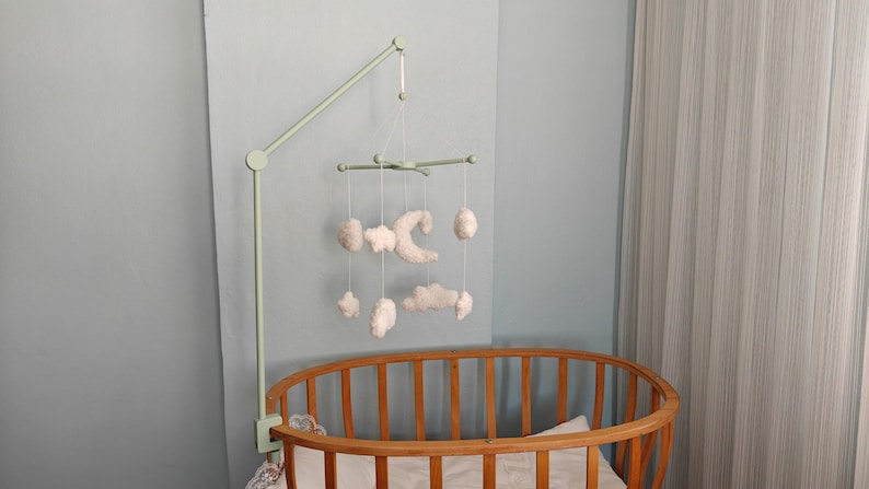 May include: A light green metal arm attached to a wooden oval crib, suspending a mobile with white plush shapes including a crescent moon, stars, and clouds hanging from thin white strings. The mobile features a central cross-shaped frame.