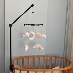 May include: A black metal mobile arm attached to a wooden crib, featuring a black crossbar from which white plush clouds and a crescent moon are suspended by thin white strings. The mobile hangs centrally over the crib's slatted wooden frame.