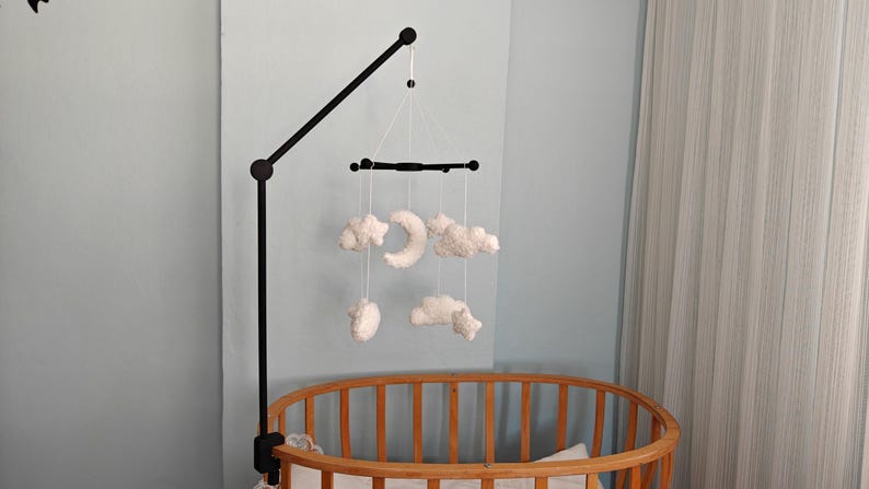 May include: A black metal mobile arm attached to a wooden crib, featuring a black crossbar from which white plush clouds and a crescent moon are suspended by thin white strings. The mobile hangs centrally over the crib's slatted wooden frame.