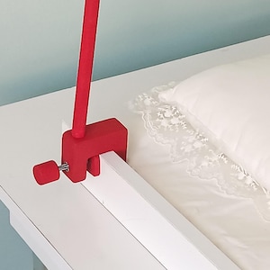 May include: A bright red C-clamp mounting bracket with a matching red tightening knob and a vertical rod attached. The clamp is secured onto the edge of a white wooden frame, featuring a simple mechanical design for attachment purposes.