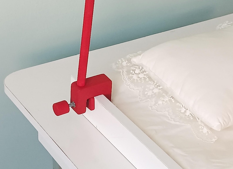 May include: A bright red C-clamp mounting bracket with a matching red tightening knob and a vertical rod attached. The clamp is secured onto the edge of a white wooden frame, featuring a simple mechanical design for attachment purposes.