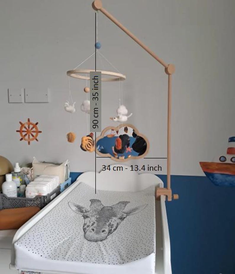 May include: A wooden baby mobile featuring felt figures of a tiger, a cloud, and a ship, suspended from a circular frame on a wooden arm. The mobile measures 35 inches in total height and 13.4 inches in width, hanging over a changing pad.