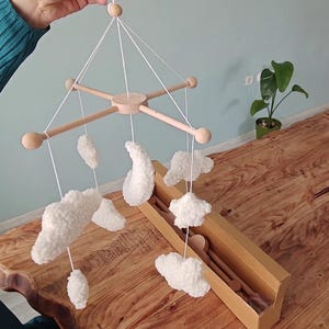 May include: A hanging mobile featuring a light-colored wooden cross frame with rounded ends. Suspended from the frame by white strings are several white, textured, plush-like shapes including clouds, a crescent moon, and a star.