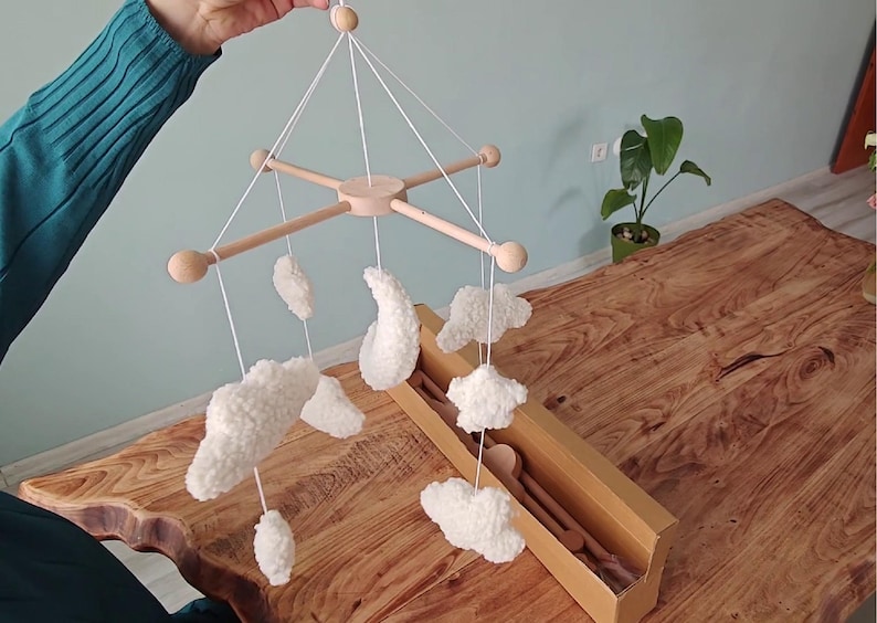 May include: A hanging mobile featuring a light-colored wooden cross frame with rounded ends. Suspended from the frame by white strings are several white, textured, plush-like shapes including clouds, a crescent moon, and a star.