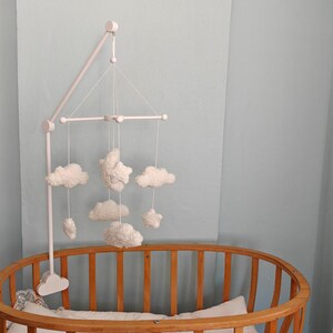 May include: A white nursery mobile featuring soft, plush cloud and star shapes suspended from a white wooden frame. The mobile is attached to a white stand that is mounted onto the side of a wooden oval crib with vertical slats.