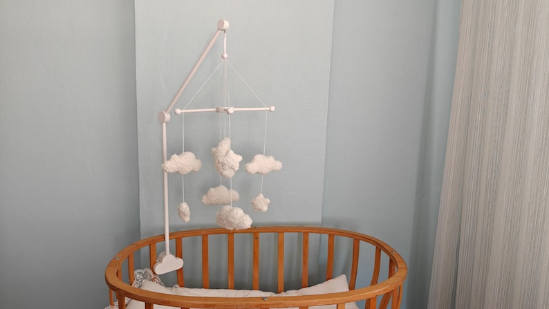 May include: A white nursery mobile featuring soft, plush cloud and star shapes suspended from a white wooden frame. The mobile is attached to a white stand that is mounted onto the side of a wooden oval crib with vertical slats.