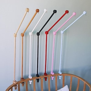 May include: A collection of seven crib mobile arms in various colors including natural wood, light brown, white, black, red, pink, and light blue. Each arm features a vertical post, a jointed angled extension, and a small metal hook at the end.