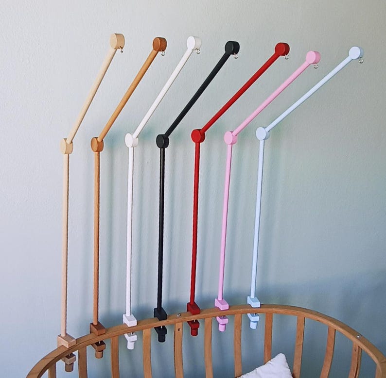 May include: A collection of seven crib mobile arms in various colors including natural wood, light brown, white, black, red, pink, and light blue. Each arm features a vertical post, a jointed angled extension, and a small metal hook at the end.