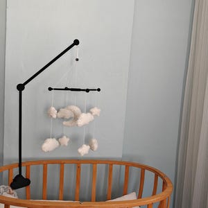 May include: A black metal mobile arm attached to a wooden crib, featuring a hanging mobile with white plush clouds, stars, and a crescent moon suspended by thin white strings from a black horizontal crossbar frame.