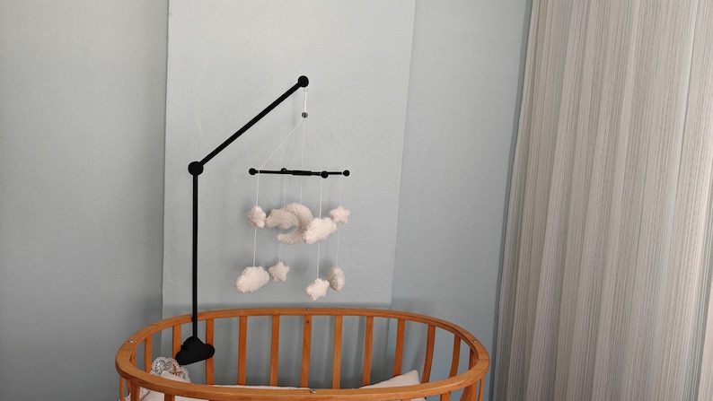 May include: A black metal mobile arm attached to a wooden crib, featuring a hanging mobile with white plush clouds, stars, and a crescent moon suspended by thin white strings from a black horizontal crossbar frame.