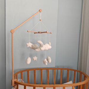 May include: A wooden crib mobile arm attached to a round wooden crib. The mobile features white, plush, celestial-themed hanging ornaments including a crescent moon, several clouds, and stars suspended by thin white strings from a wooden crossbar.