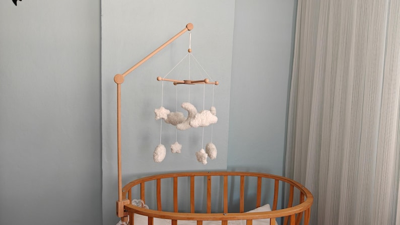 May include: A wooden crib mobile arm attached to a round wooden crib. The mobile features white, plush, celestial-themed hanging ornaments including a crescent moon, several clouds, and stars suspended by thin white strings from a wooden crossbar.