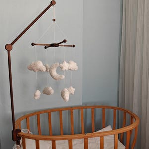 May include: A wooden crib mobile arm holds a hanging mobile featuring white plush clouds, stars, and a crescent moon suspended by thin white strings from a dark brown wooden cross-shaped frame, positioned above a wooden oval-shaped crib.