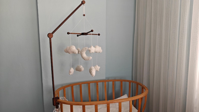 May include: A wooden crib mobile arm holds a hanging mobile featuring white plush clouds, stars, and a crescent moon suspended by thin white strings from a dark brown wooden cross-shaped frame, positioned above a wooden oval-shaped crib.