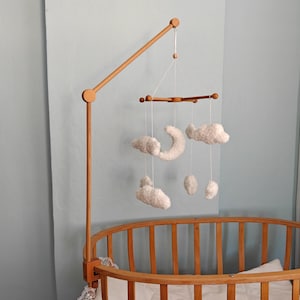 May include: A wooden crib mobile arm attached to an oval wooden crib. The mobile features white, plush, cloud-shaped and crescent moon-shaped ornaments suspended by thin white strings from a horizontal wooden crossbar frame.