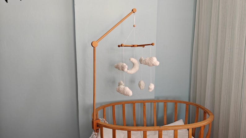 May include: A wooden crib mobile arm attached to an oval wooden crib. The mobile features white, plush, cloud-shaped and crescent moon-shaped ornaments suspended by thin white strings from a horizontal wooden crossbar frame.