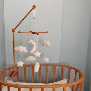 May include: A wooden crib mobile arm attached to an oval wooden crib, featuring hanging white plush ornaments shaped like clouds, stars, and a crescent moon suspended by thin white strings from a central wooden cross-shaped frame structure.