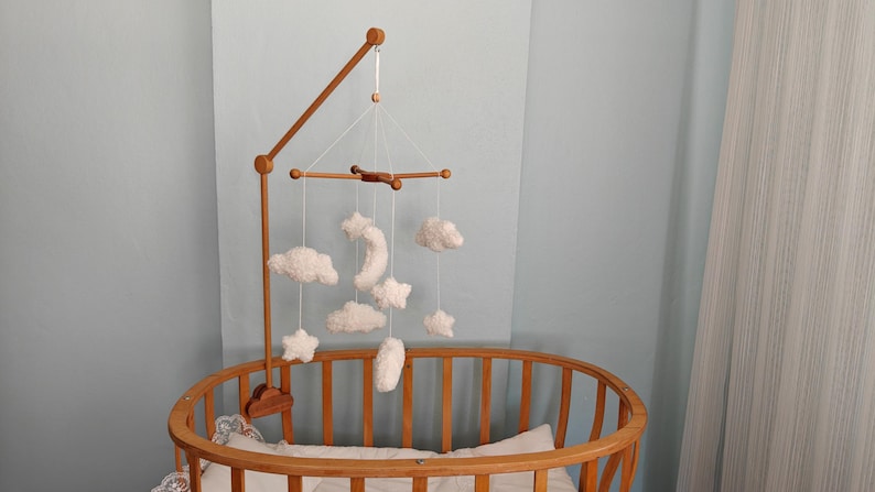 May include: A wooden crib mobile arm attached to an oval wooden crib, featuring hanging white plush ornaments shaped like clouds, stars, and a crescent moon suspended by thin white strings from a central wooden cross-shaped frame structure.