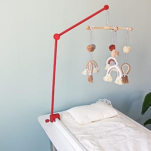 May include: A red metal mobile arm clamp attached to a white surface, featuring a wooden crossbar from which hang several white, brown, and tan yarn pom-poms and small macrame rainbow shapes with fringe details, suspended above a white pillow.