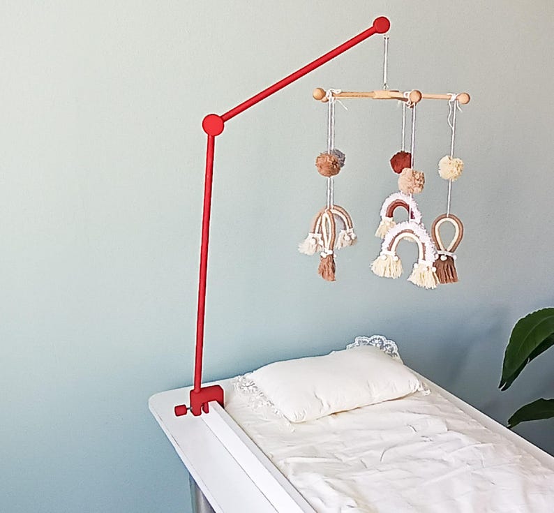 May include: A red metal mobile arm clamp attached to a white surface, featuring a wooden crossbar from which hang several white, brown, and tan yarn pom-poms and small macrame rainbow shapes with fringe details, suspended above a white pillow.