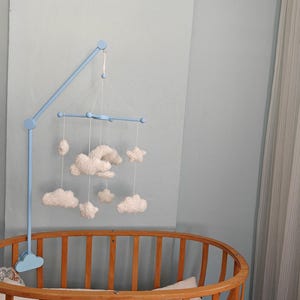 May include: A light blue wooden mobile arm attached to a crib, featuring a light blue crossbar from which several white, soft, plush-textured clouds and a star are suspended by thin white strings, creating a hanging decorative arrangement.