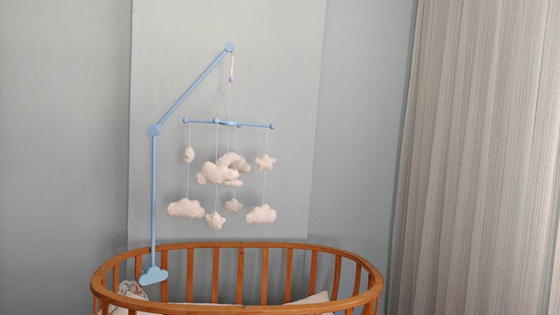 May include: A light blue wooden mobile arm attached to a crib, featuring a light blue crossbar from which several white, soft, plush-textured clouds and a star are suspended by thin white strings, creating a hanging decorative arrangement.