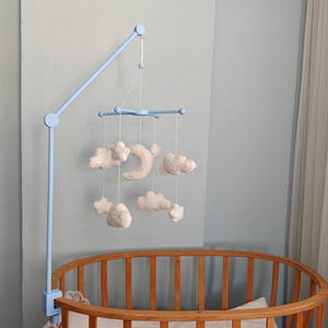 May include: A light blue mobile arm attached to a wooden crib, featuring hanging white plush clouds and a crescent moon suspended by thin white strings from a central blue cross-shaped frame, creating a soft, layered celestial arrangement.