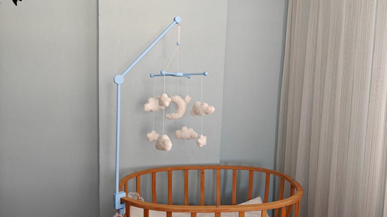 May include: A light blue mobile arm attached to a wooden crib, featuring hanging white plush clouds and a crescent moon suspended by thin white strings from a central blue cross-shaped frame, creating a soft, layered celestial arrangement.
