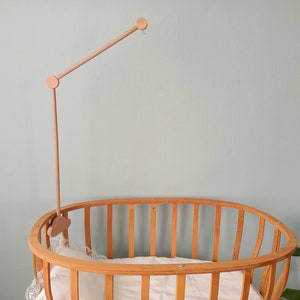 May include: A wooden baby crib with a natural finish and a wooden mobile arm attached to the top. The crib has a white mattress and a white sheet.