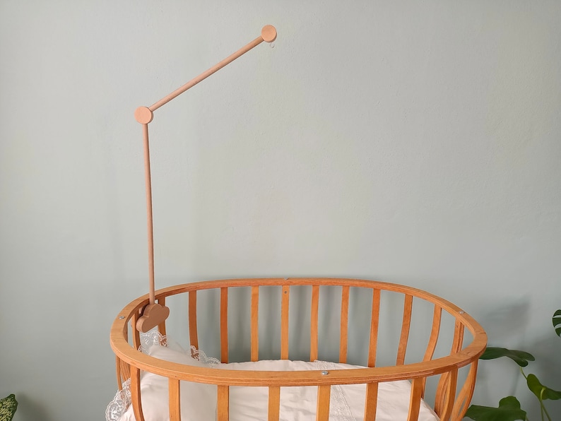 May include: A wooden baby crib with a natural finish and a wooden mobile arm attached to the top. The crib has a white mattress and a white sheet.