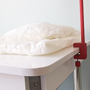 May include: A white quilted mattress pad with a decorative lace-trimmed pillow resting on top, secured to a white table surface by a red metal clamp mount with a vertical red pole extending upward from the side of the table edge.